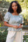 woman wearing a light blue puff sleeve top with lace detailing and ruffled hem, posing outdoors among greenery