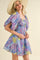 woman wearing a floral purple mini dress with puff sleeves and smocked waist, styled with a wide-brimmed hat
