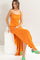 woman wearing an orange spaghetti strap maxi dress and green high heeled sandals, sitting on a white pedestal