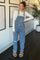 woman wearing light blue denim overalls with ripped details and a long sleeve gray top, styled with fuzzy slides