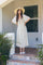woman wearing a floral midi dress with short sleeves and a tie front, standing on a tiled porch with a wide-brimmed hat