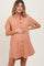 woman wearing a rust-colored maternity button-up dress with short sleeves and a gathered waist, holding her baby bump