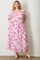 woman wearing a floral print maxi dress in pink and white with short sleeves and pockets, standing against a white wall