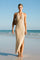 woman wearing a beige halter neck maxi dress with a tie detail and side slit, standing on the beach