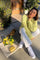 woman wearing a pastel yellow cable-knit sweater and white wide-leg pants, sitting on the ground next to a tote bag and flowers