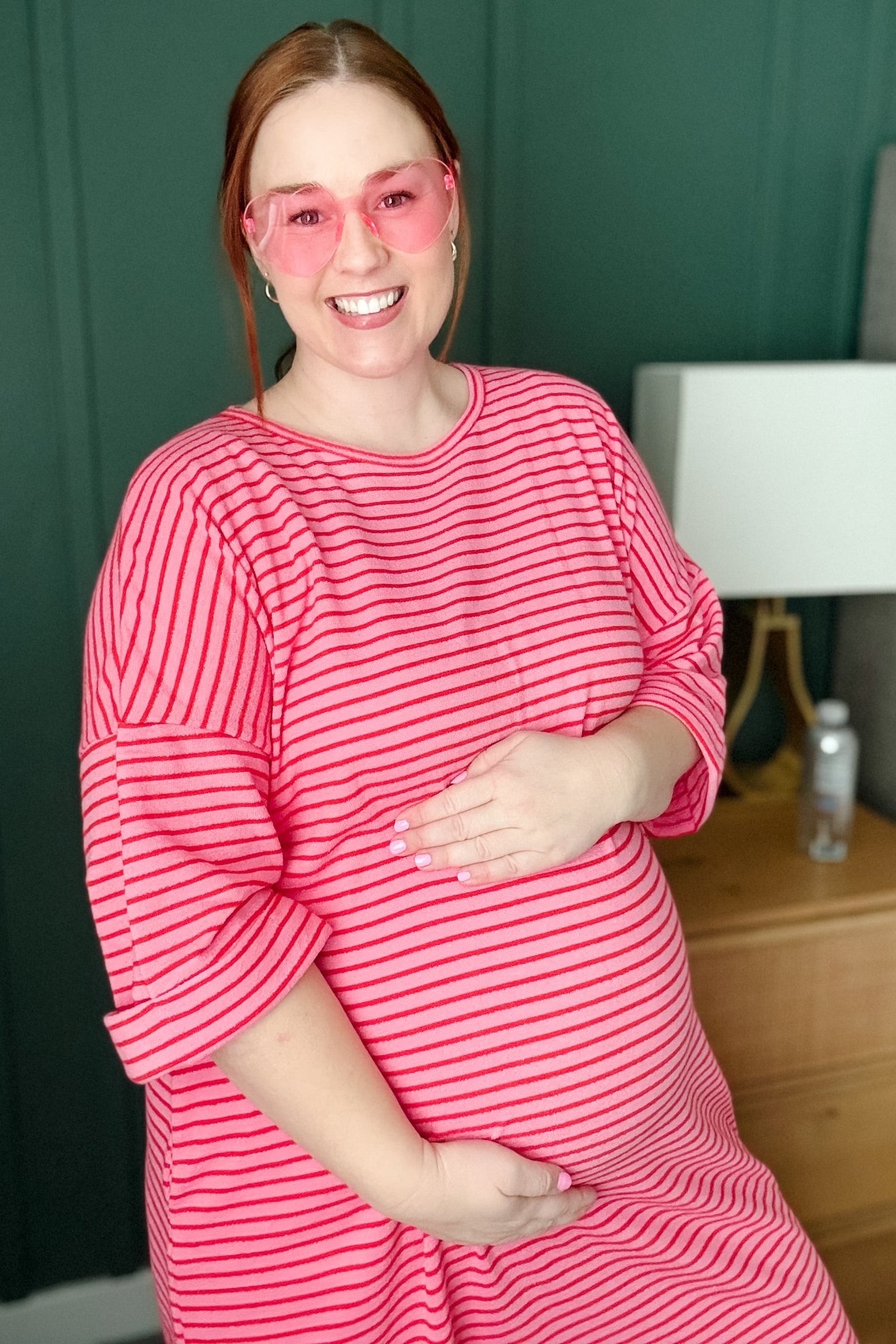 woman wearing a pink and white striped maternity dress and holding her baby bump while smiling