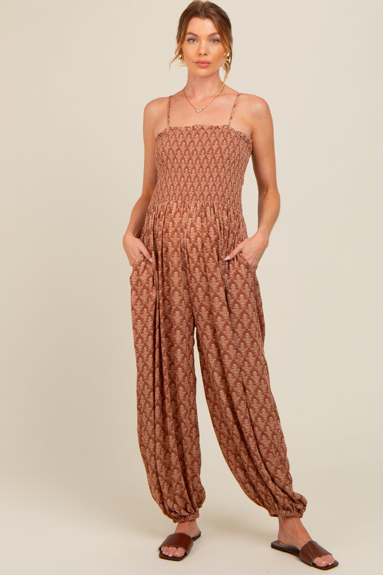 woman wearing a rust patterned maternity jumpsuit with adjustable straps and pockets, showing a baby bump