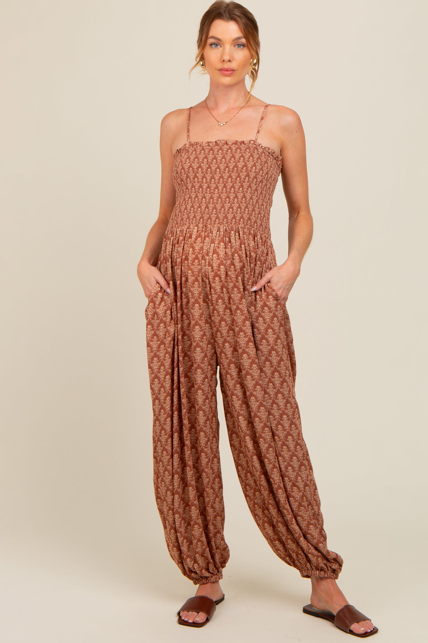 woman wearing a rust patterned maternity jumpsuit with adjustable straps and pockets, showing a baby bump