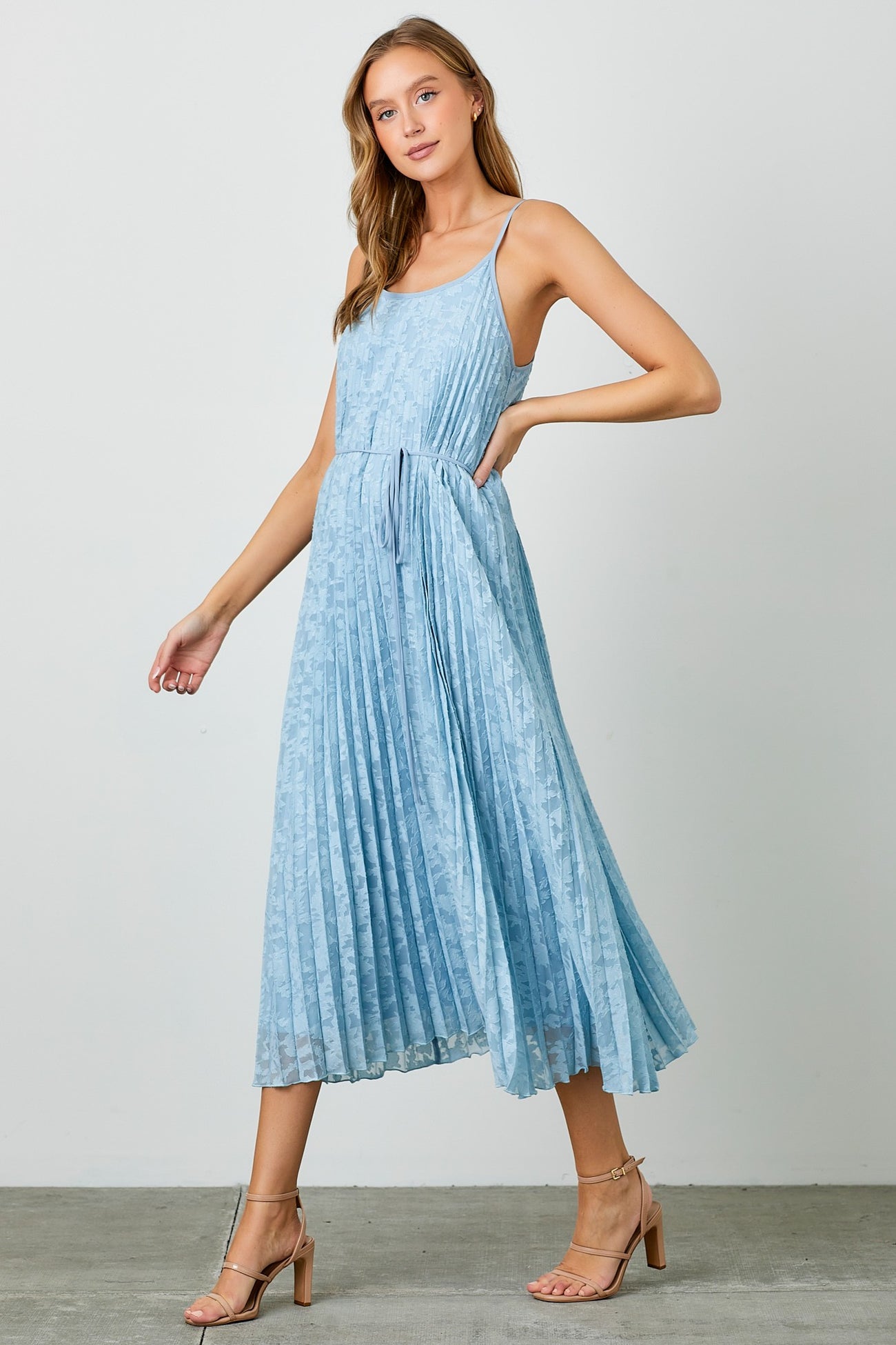 woman wearing a light blue sleeveless pleated maxi dress with a cinched waist and strappy heels
