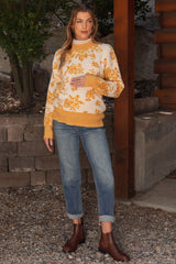 woman wearing a cream and mustard floral maternity sweater and blue jeans, holding her baby bump in an outdoor setting