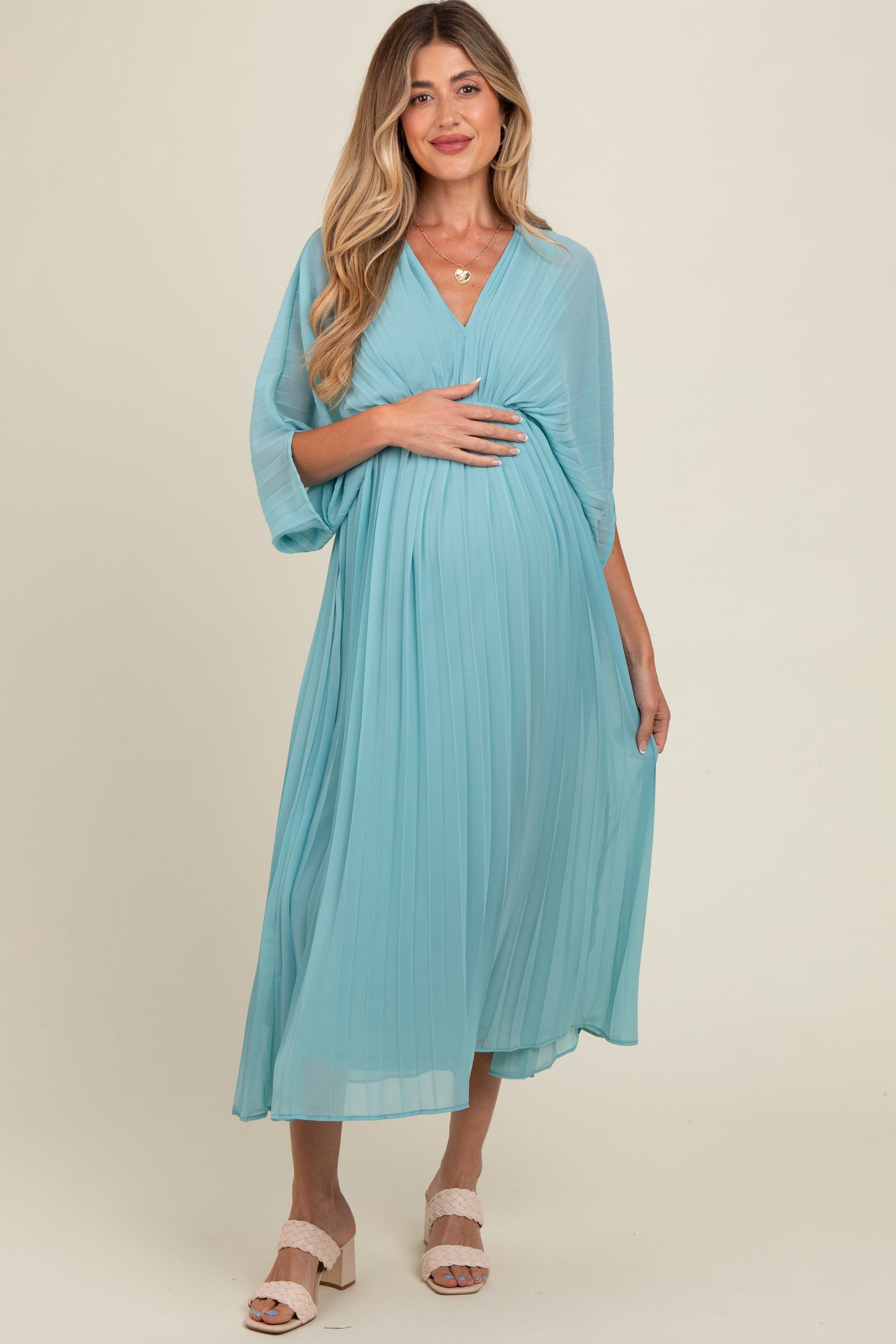 woman wearing a light blue pleated maternity dress with a v-neck and draped sleeves, cradling her baby bump