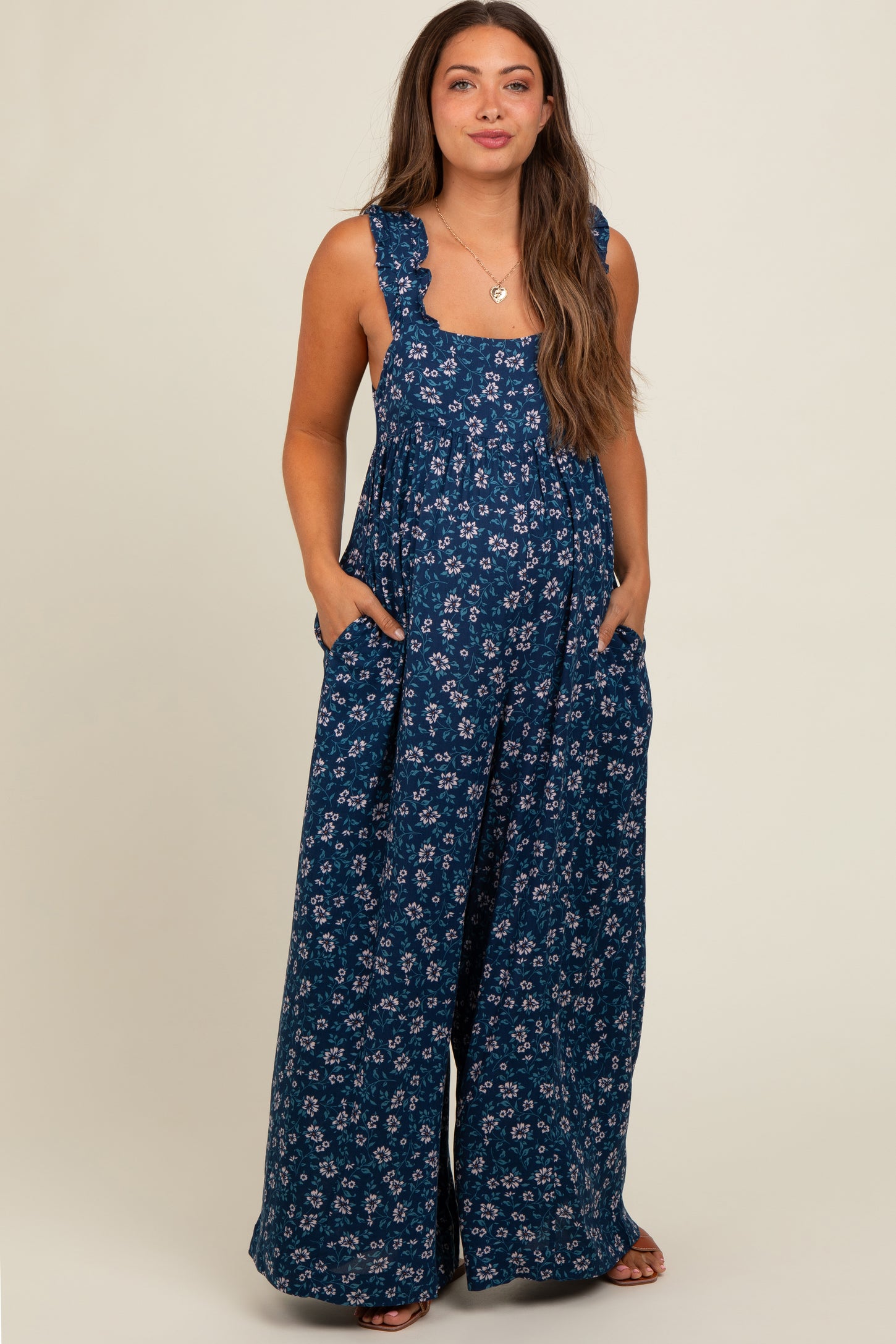 woman wearing a navy blue floral print maternity jumpsuit with pockets and ruffled straps, showcasing her baby bump