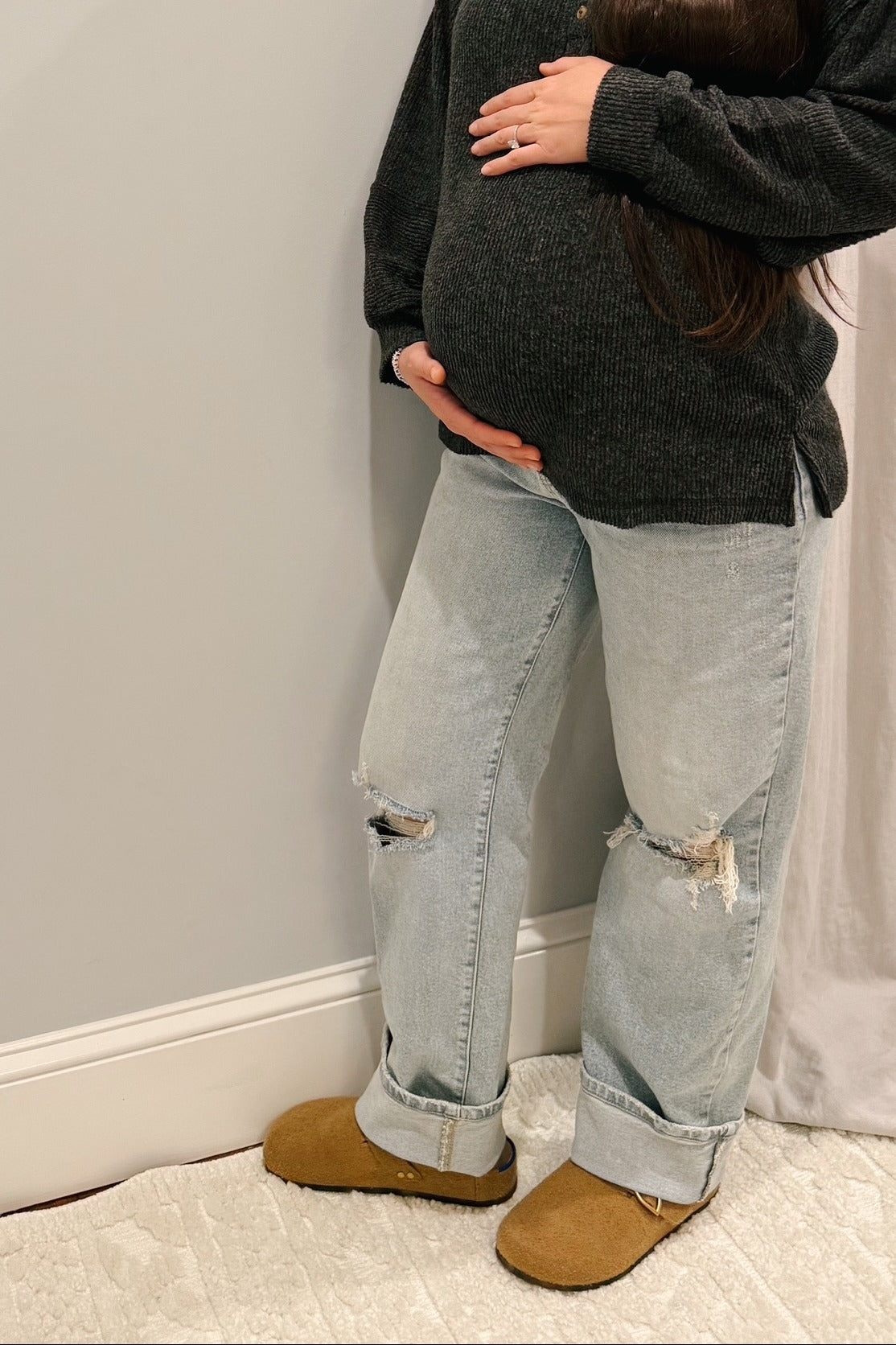 woman wearing a dark gray sweater and distressed light blue maternity jeans, cradling her baby bump