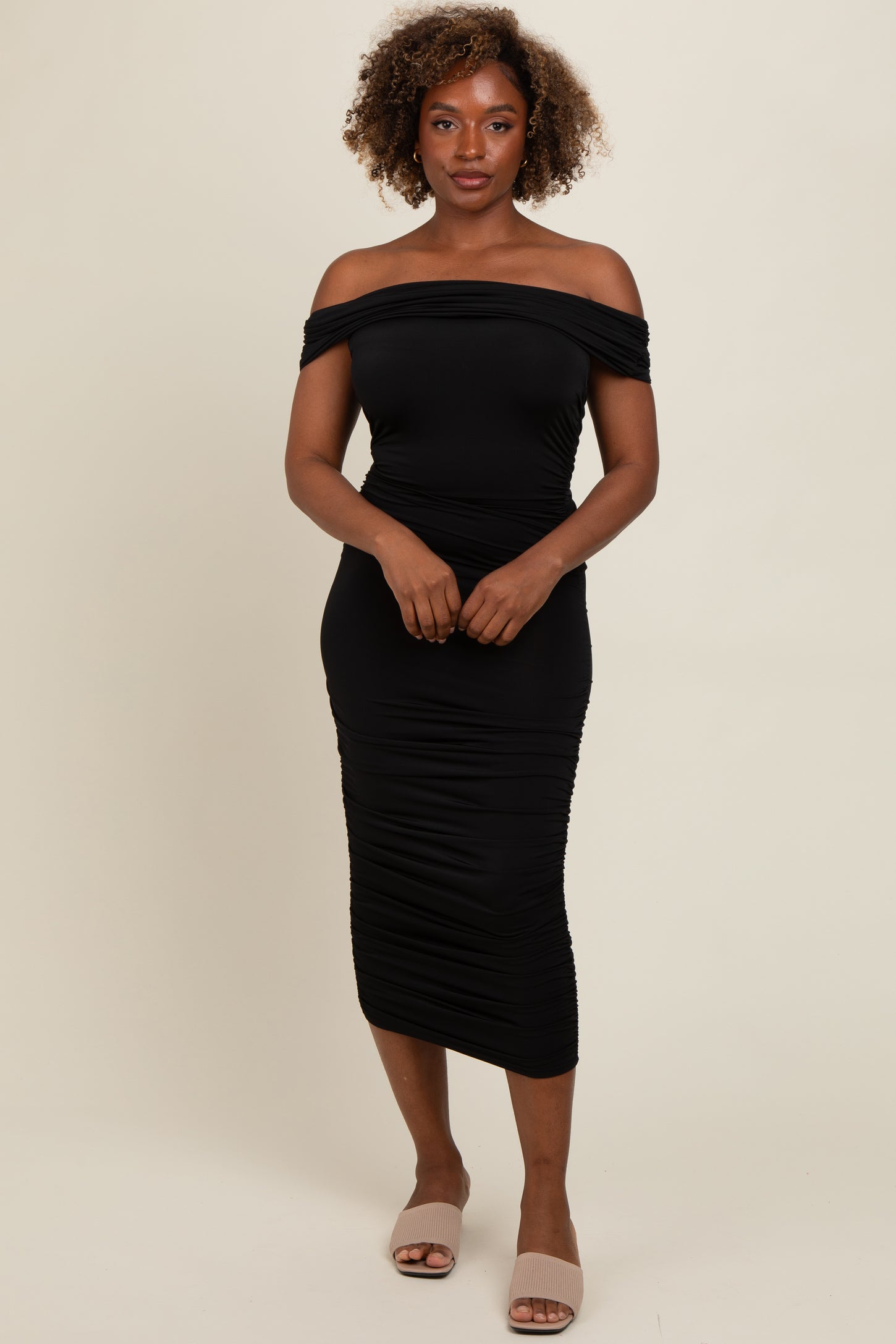 woman wearing a black off-shoulder bodycon midi dress with ruching details and slip-on beige sandals