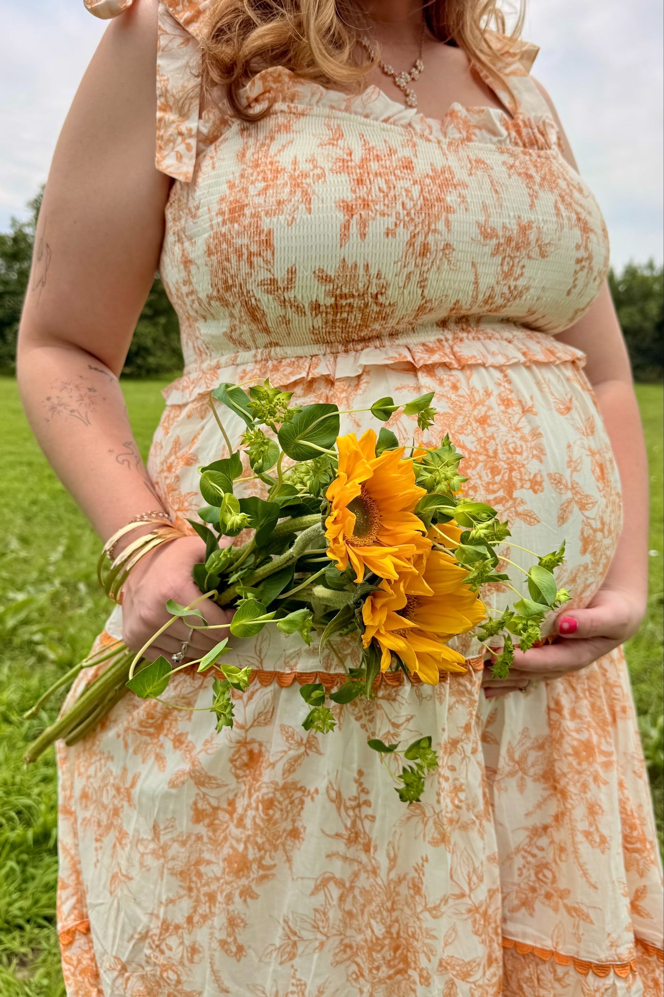woman wearing a floral orange and cream maternity dress holding a bouquet of sunflowers with a visible baby bump