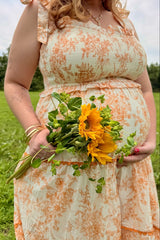 woman wearing a floral orange and cream maternity dress holding a bouquet of sunflowers with a visible baby bump