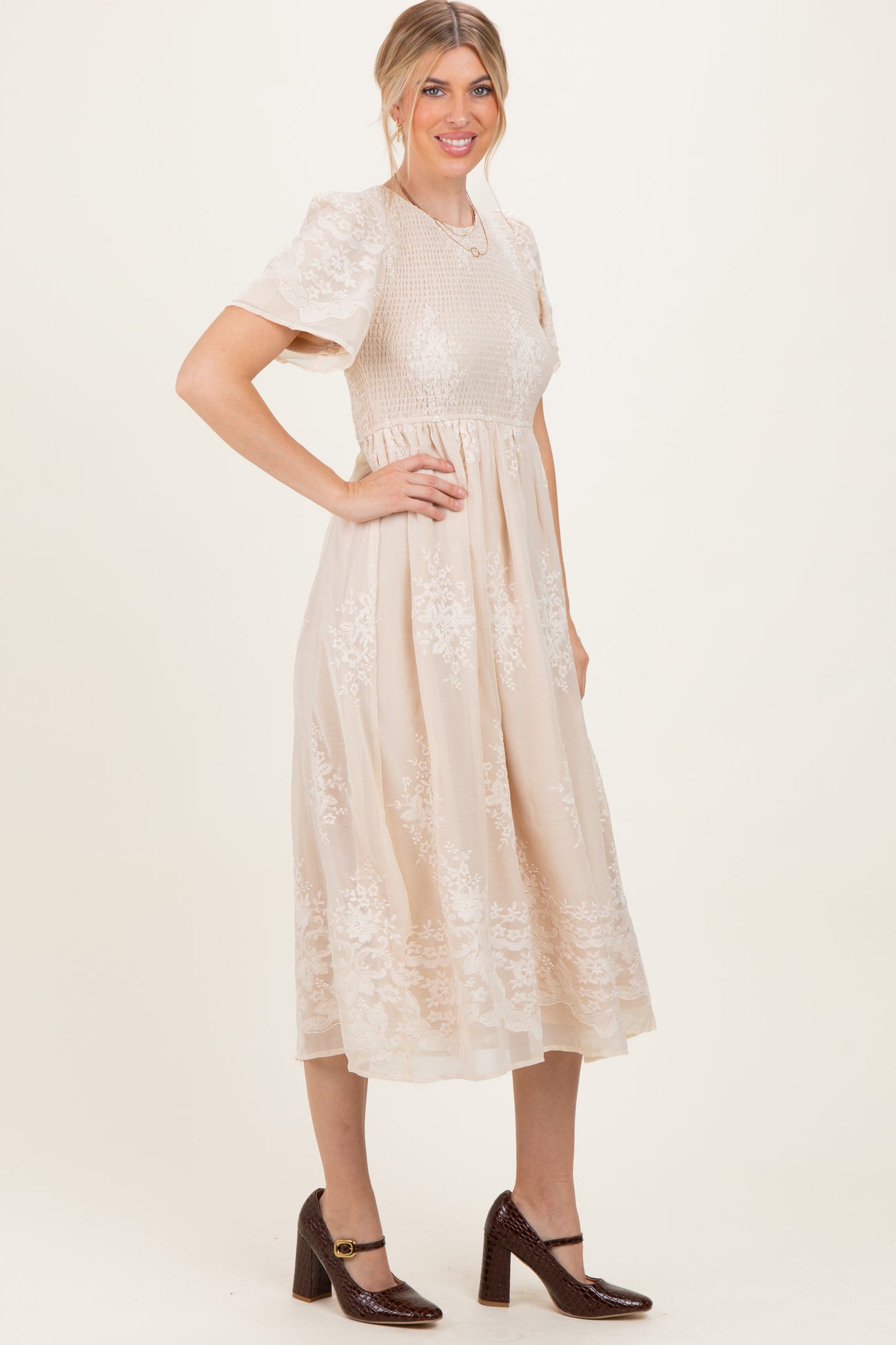 woman wearing a beige midi dress with floral embroidery and short sleeves, standing with one hand on her hip