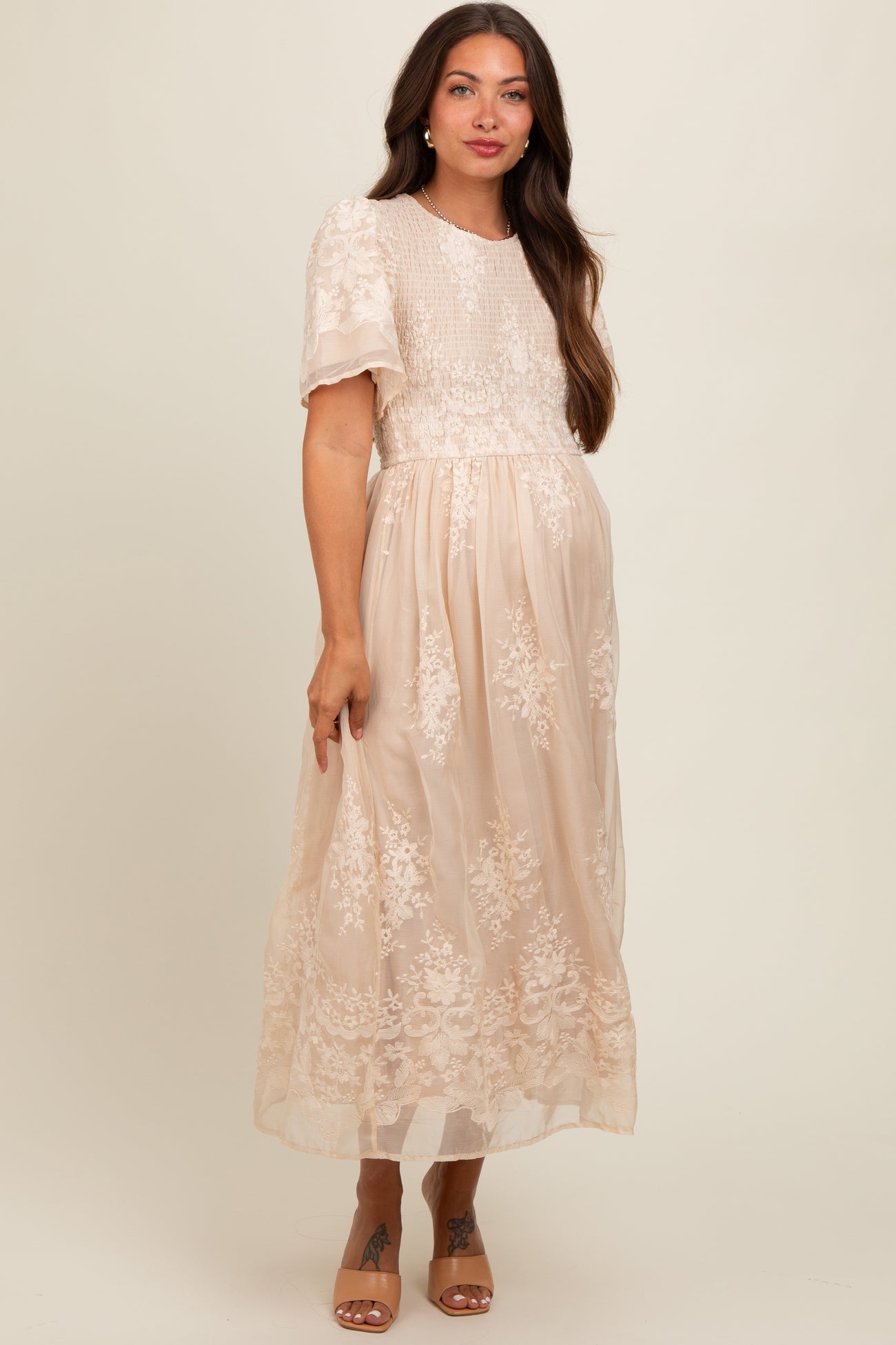 woman wearing a cream embroidered maternity maxi dress with short puff sleeves and a flowing skirt, highlighting her baby bump