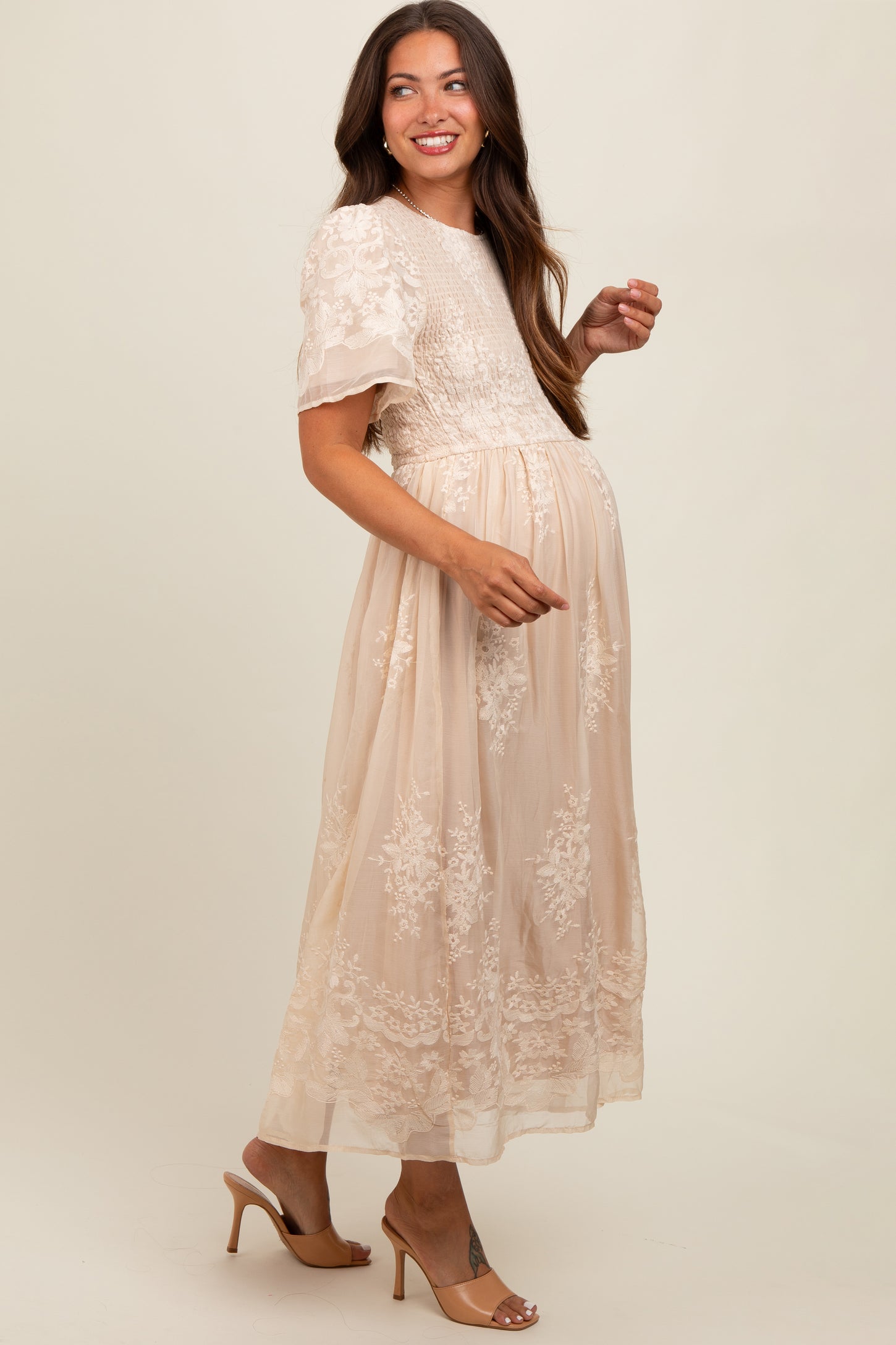 woman wearing a nude embroidered maternity dress with short sleeves and a flowing skirt, smiling and showing her baby bump