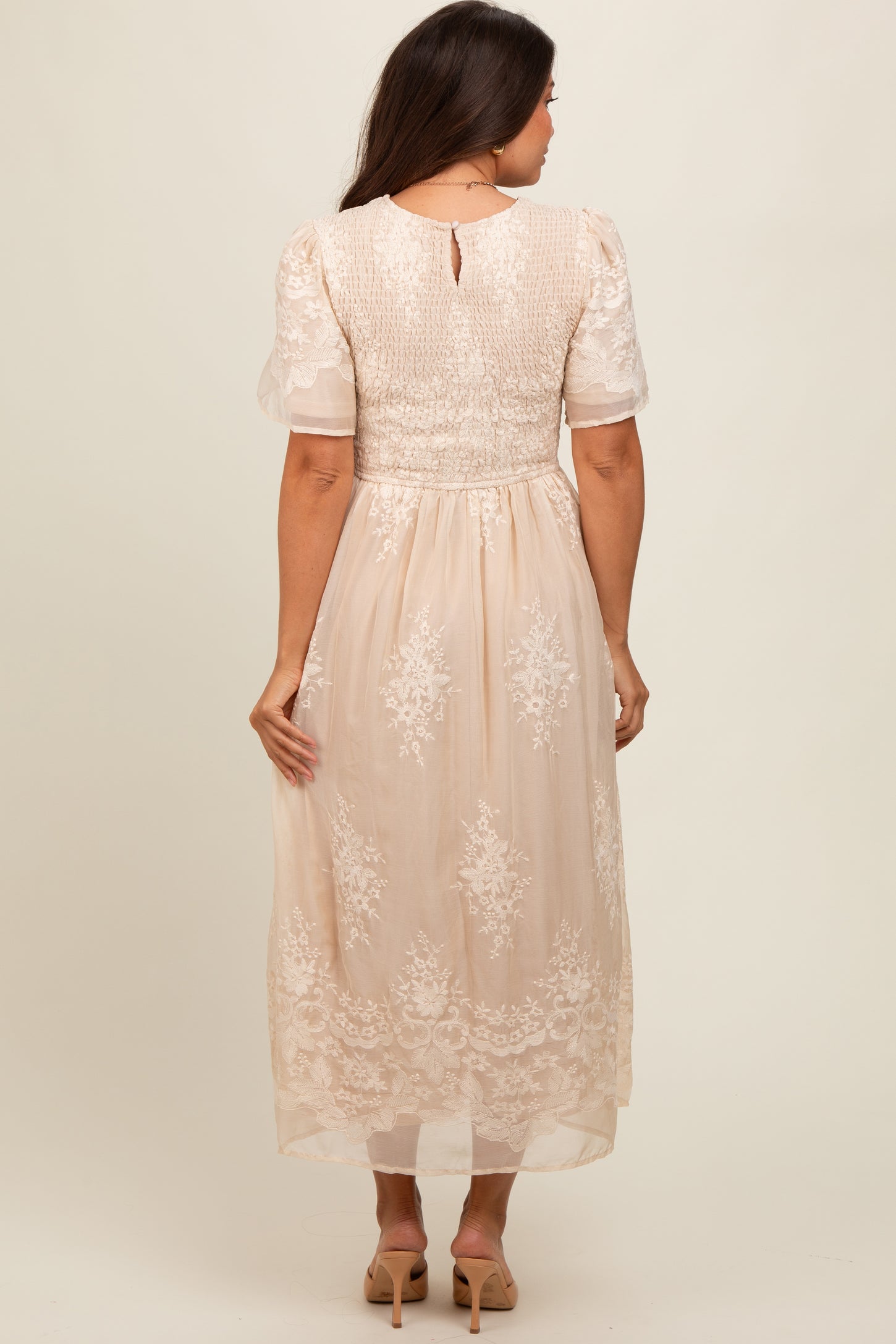 woman wearing a beige maternity dress with sheer sleeves and floral embroidery, showing the back view