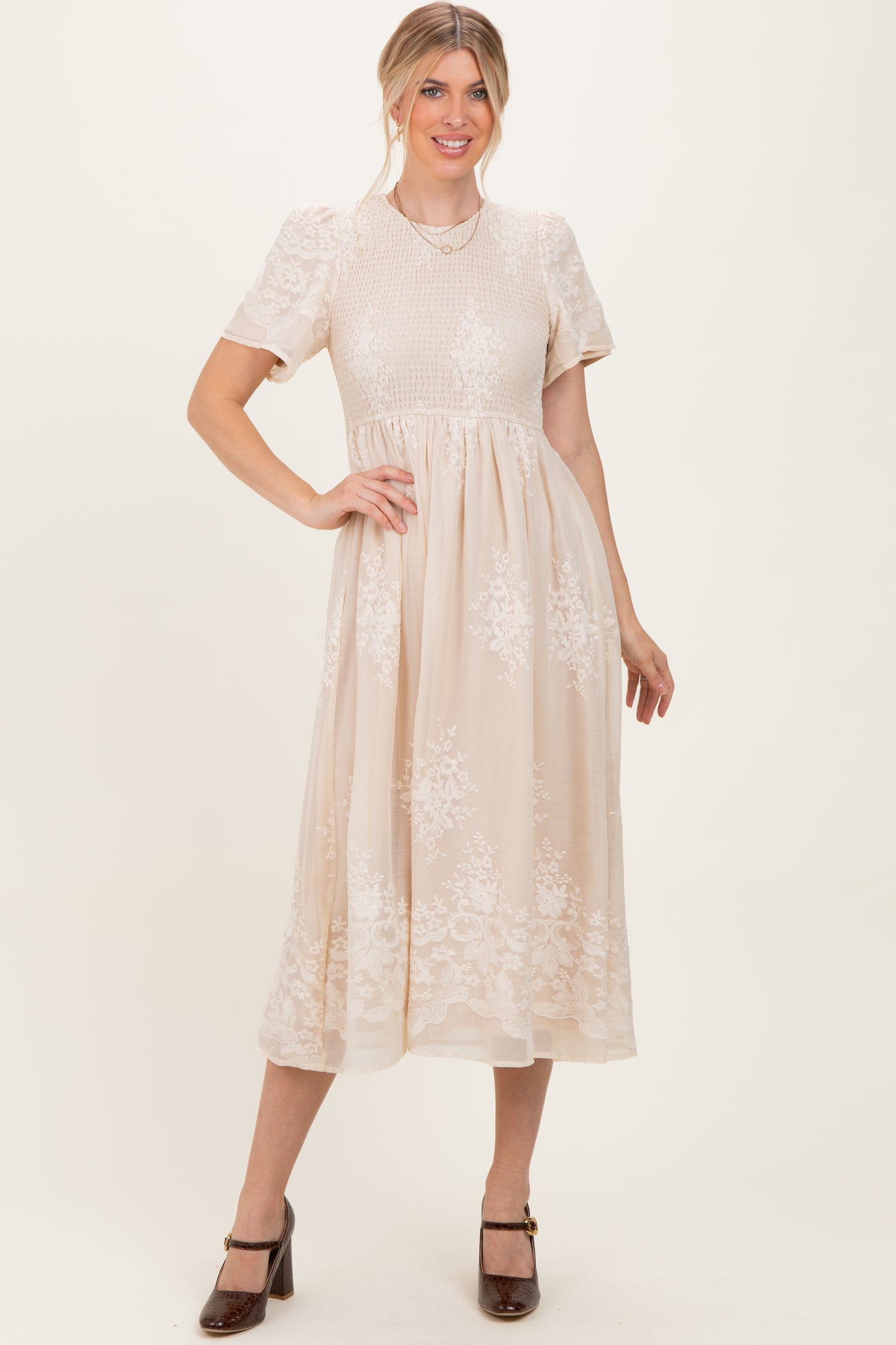 woman wearing a beige midi dress with short sleeves and floral embroidery, posing with one hand on her hip