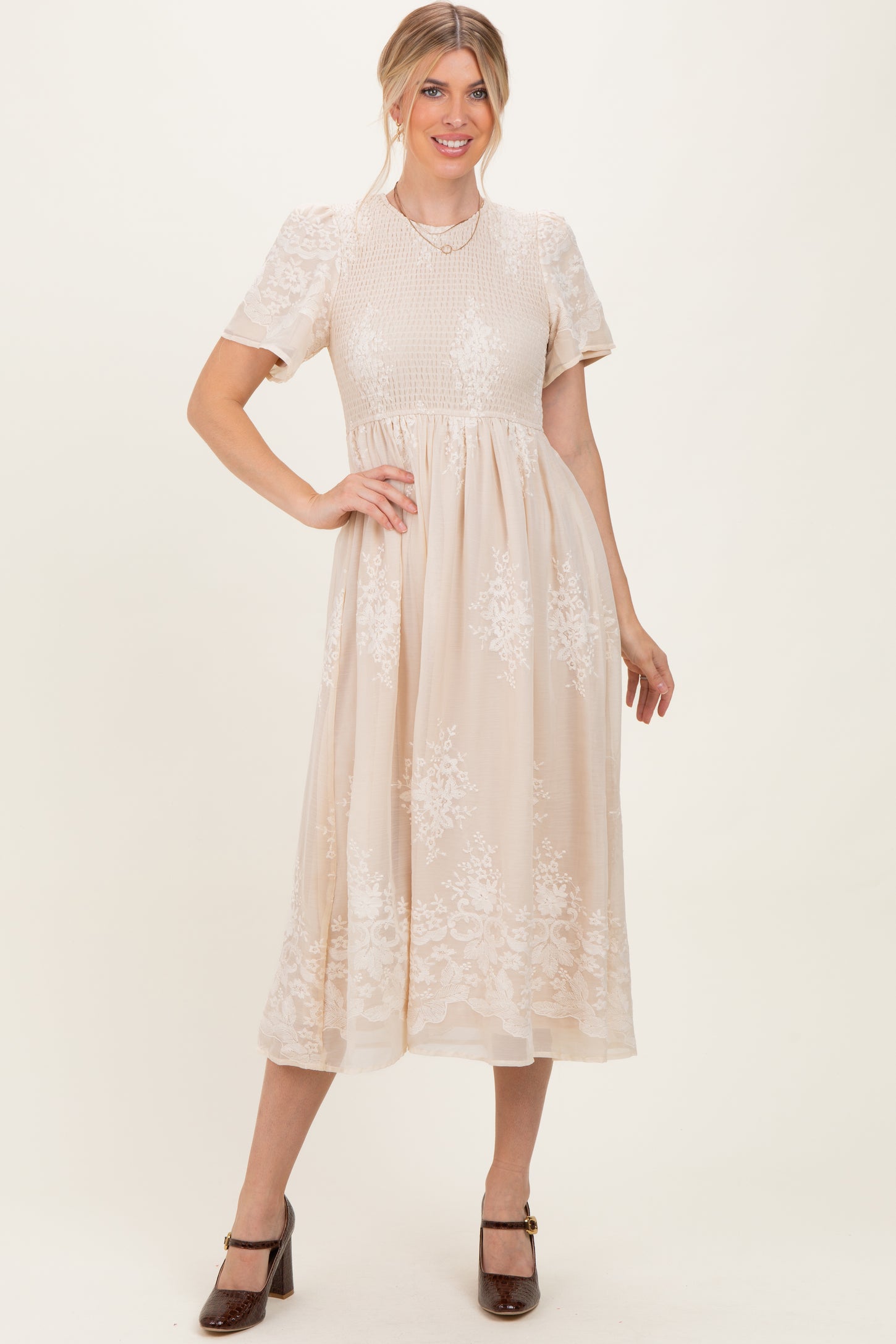 woman wearing a beige midi dress with short sleeves and floral embroidery, posing with one hand on her hip