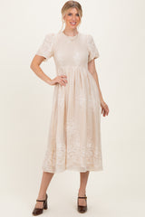 woman wearing a beige midi dress with short sleeves and floral embroidery, posing with one hand on her hip