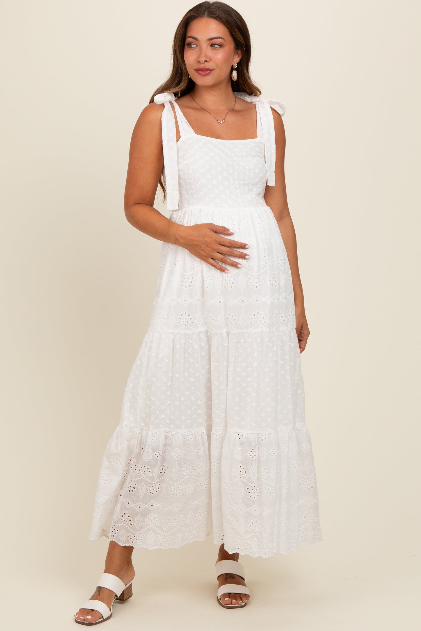 woman wearing a white maternity maxi dress with lace details and tie straps, holding her baby bump