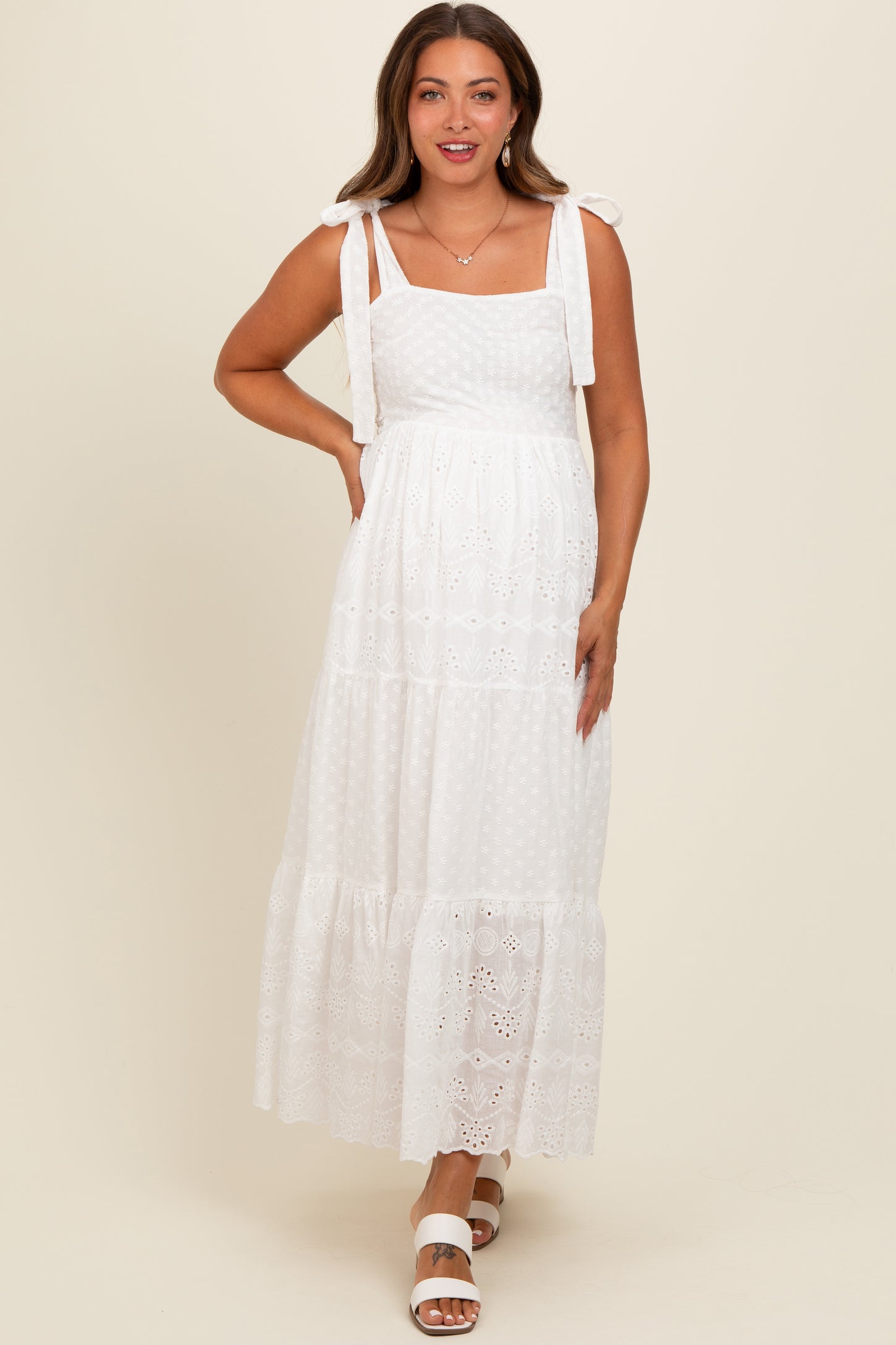 woman wearing a white maternity maxi dress with shoulder ties and lace details, smiling and showing a baby bump