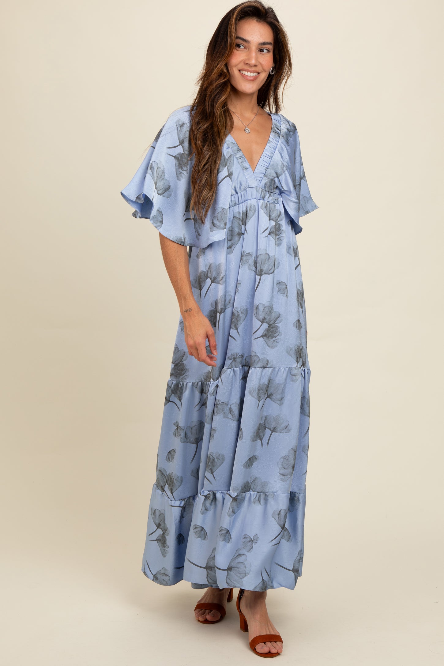 woman wearing a light blue floral maxi dress with flutter sleeves and a tiered skirt, smiling against a neutral background