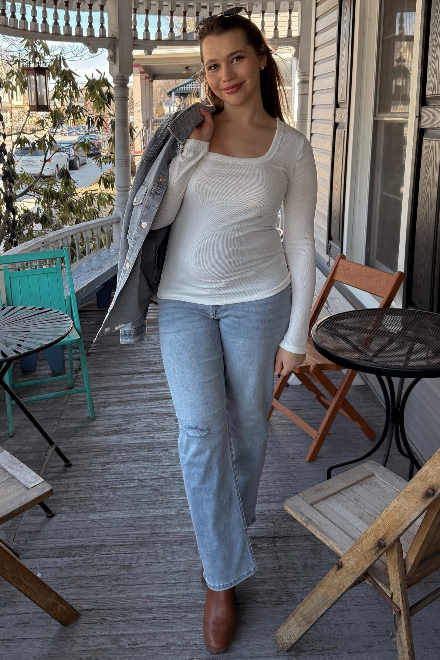 woman wearing a white long sleeve maternity top and light blue jeans, holding a denim jacket over her shoulder