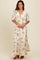 woman wearing a cream floral maxi dress with short sleeves and a ruffled hem, standing and smiling