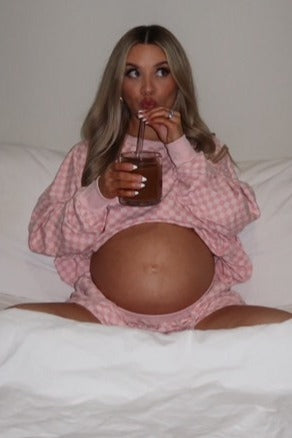 woman wearing a pink checkered maternity loungewear set with a visible baby bump, sipping a drink while sitting on a bed