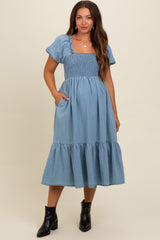 woman wearing a light blue maternity dress with short sleeves, smocked bodice, and ruffled hem, posing with hands in pockets