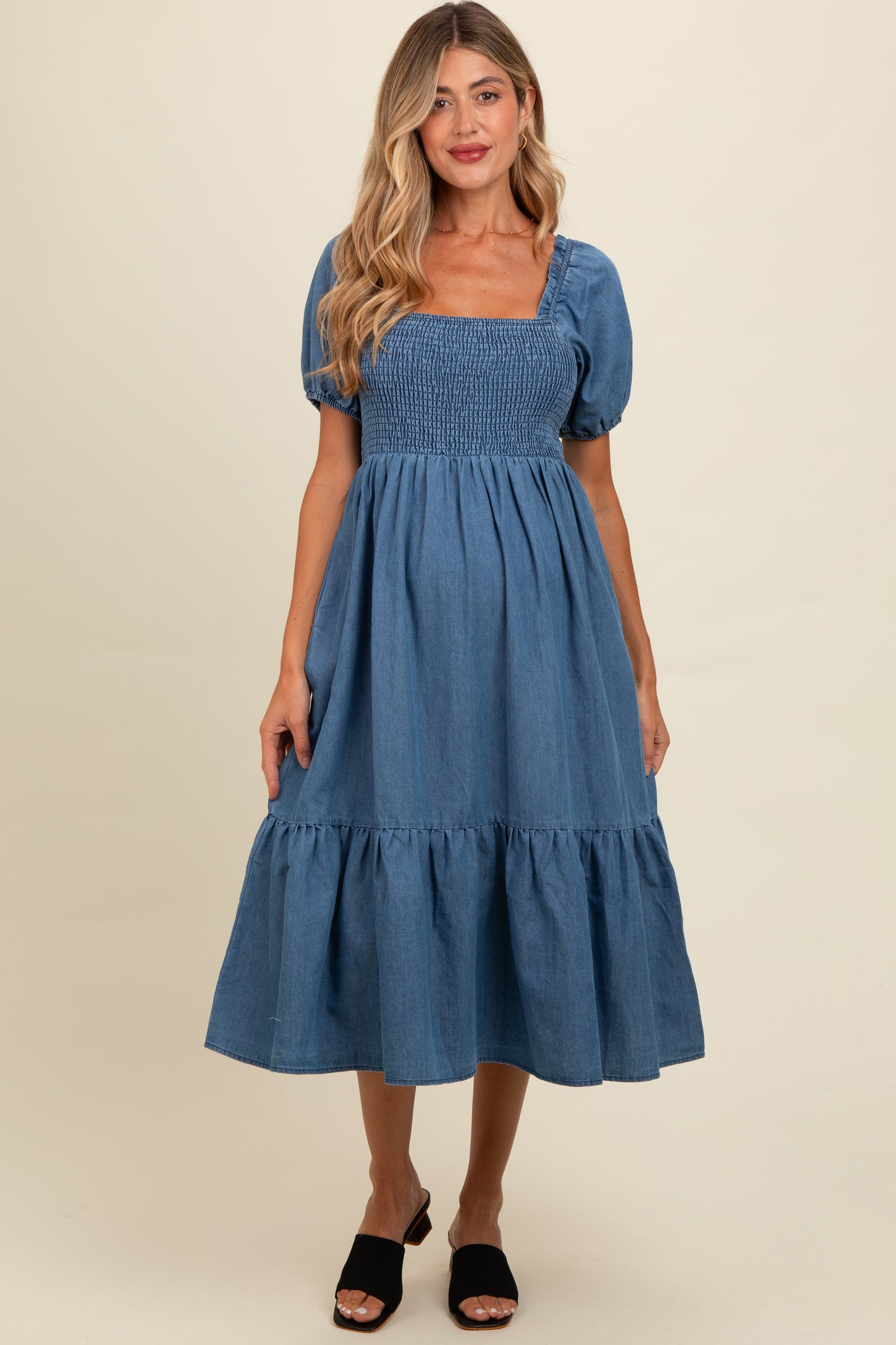 woman wearing a denim blue maternity dress with puff sleeves and a tiered hem, smiling at the camera