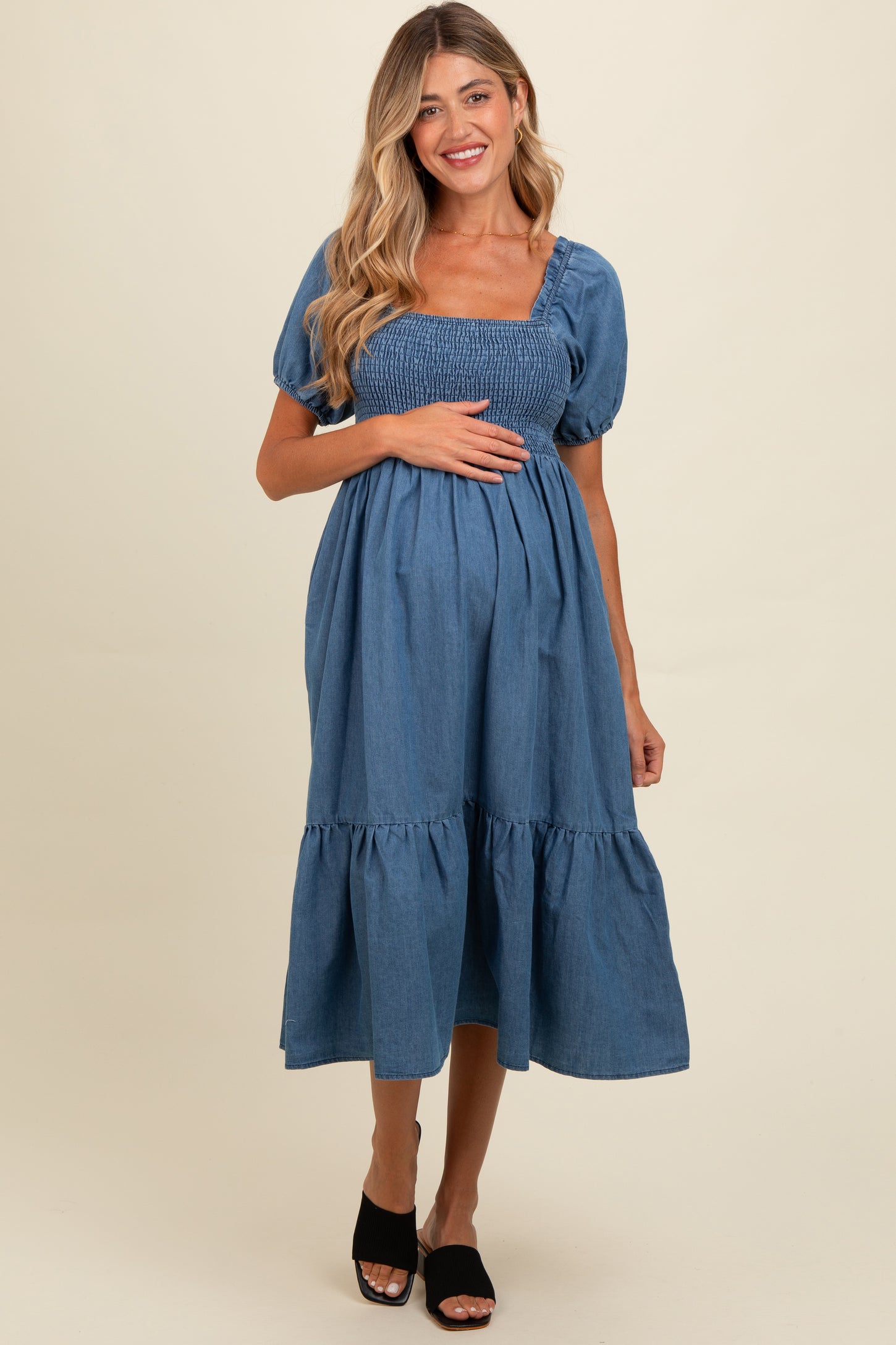 woman wearing a blue denim maternity dress with puff sleeves and a tiered skirt, holding her baby bump