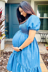 woman wearing a blue denim maternity dress with puff sleeves and a gathered bodice, gently cradling her baby bump