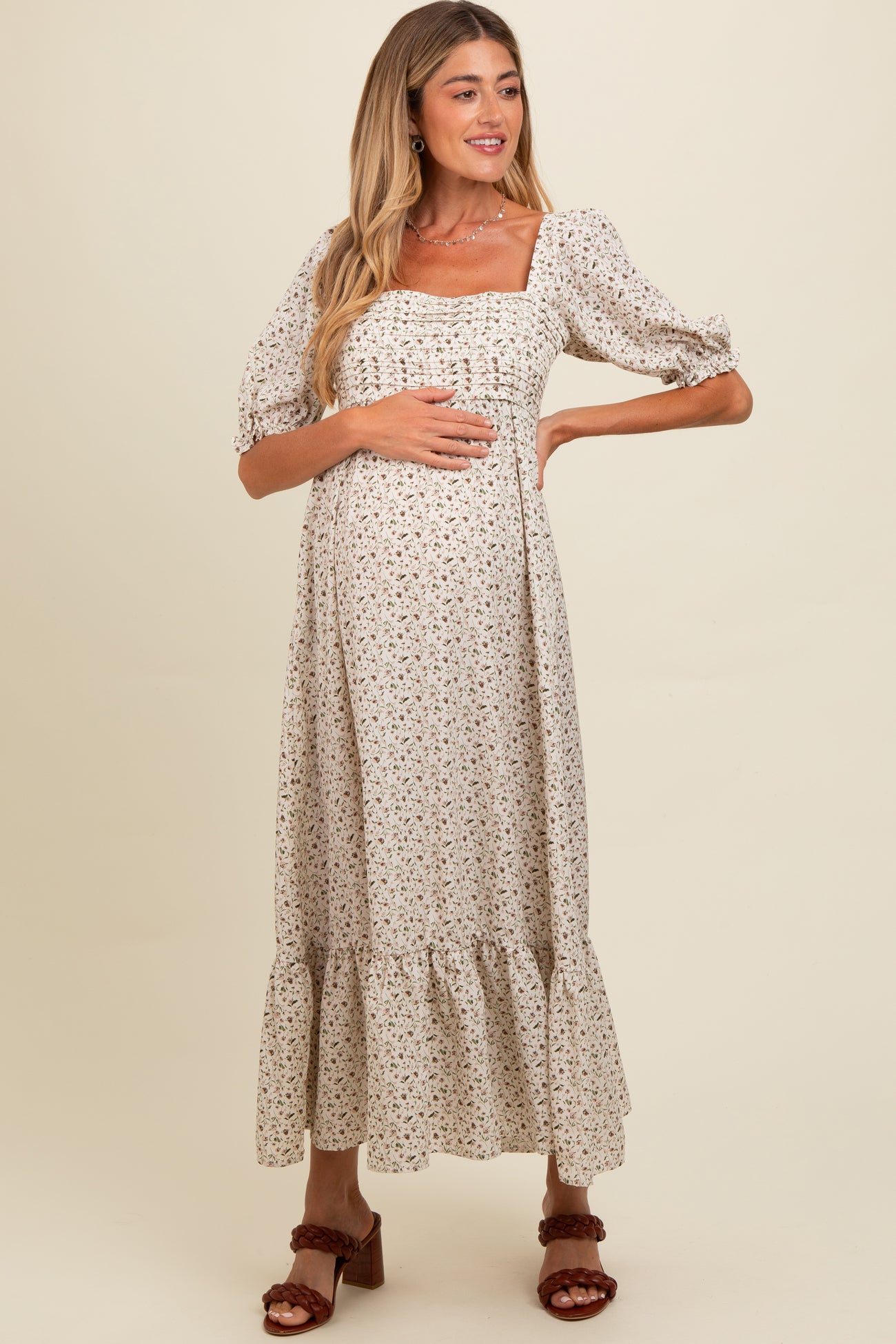 woman wearing a cream floral maternity dress with short puff sleeves and a tiered maxi skirt, holding her baby bump