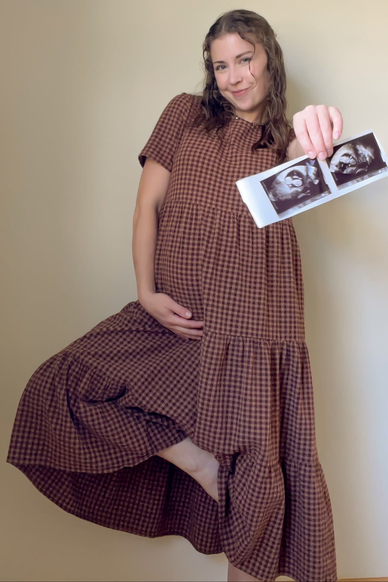 woman wearing a brown gingham maternity dress holding ultrasound photos and displaying her baby bump