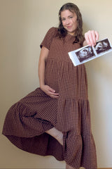 woman wearing a brown gingham maternity dress holding ultrasound photos and displaying her baby bump