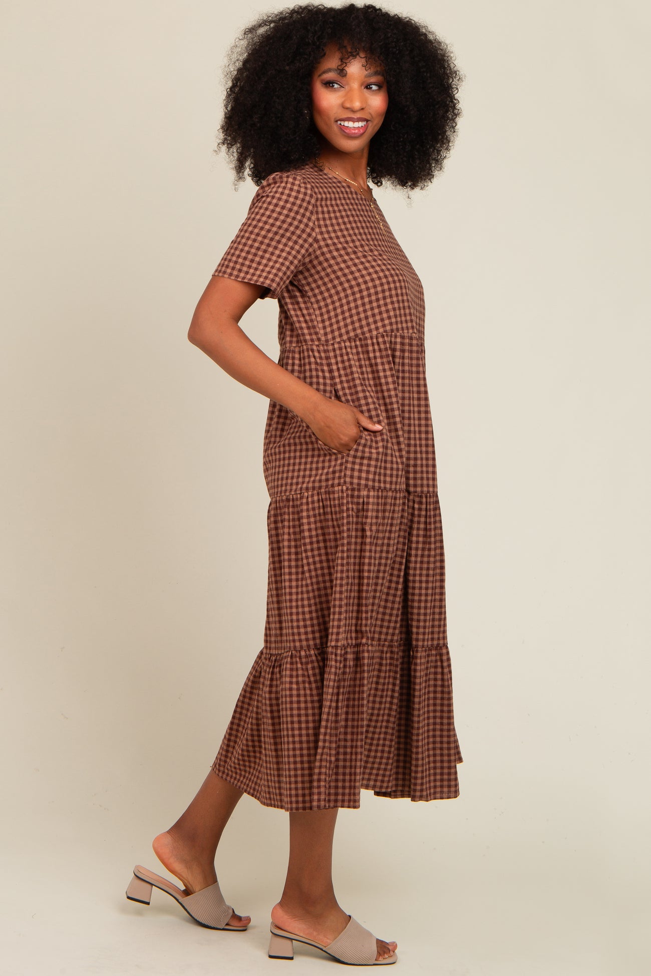 woman wearing a brown checked midi dress with short sleeves and tiered hem, standing sideways with hands in pockets