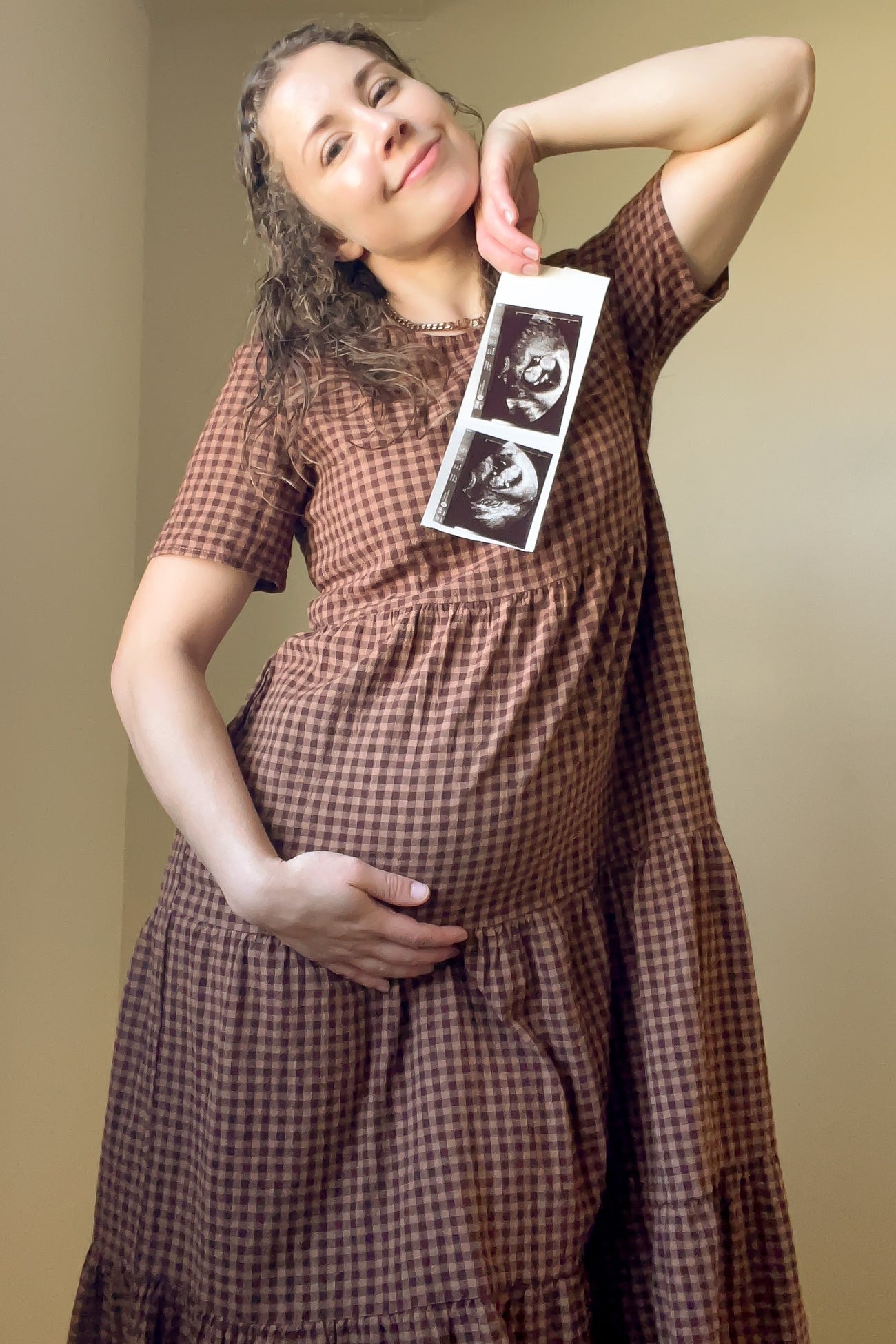 woman wearing a brown gingham maternity dress and holding an ultrasound picture next to her baby bump