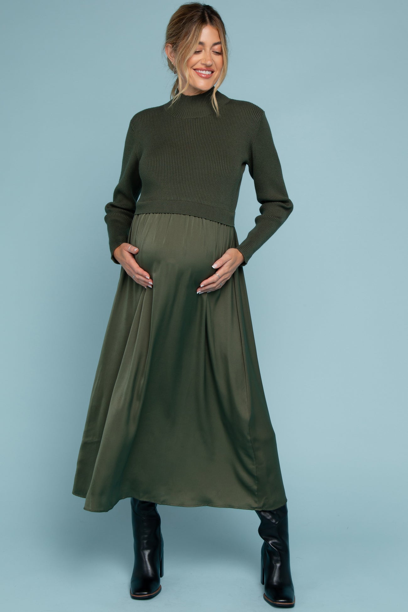 woman wearing a dark green maternity dress with a knit top and a flowy satin skirt, cradling her baby bump