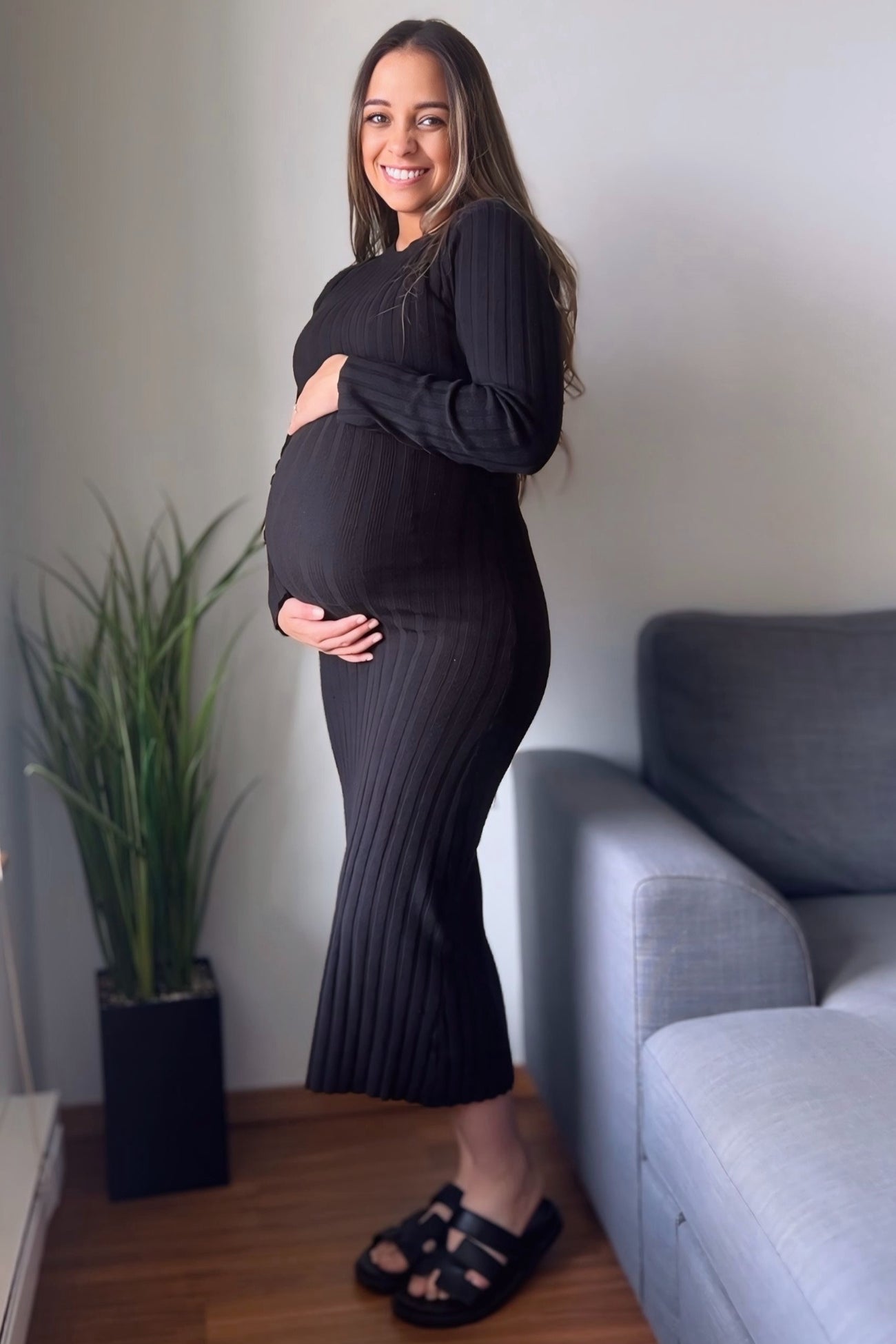 woman wearing a black ribbed maternity dress and showing her baby bump while standing next to a plant