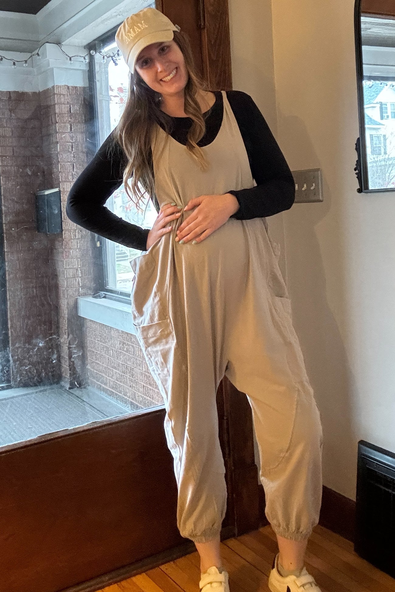 woman wearing a beige maternity jumpsuit with pockets and a baby bump, holding her belly, and a cap on her head