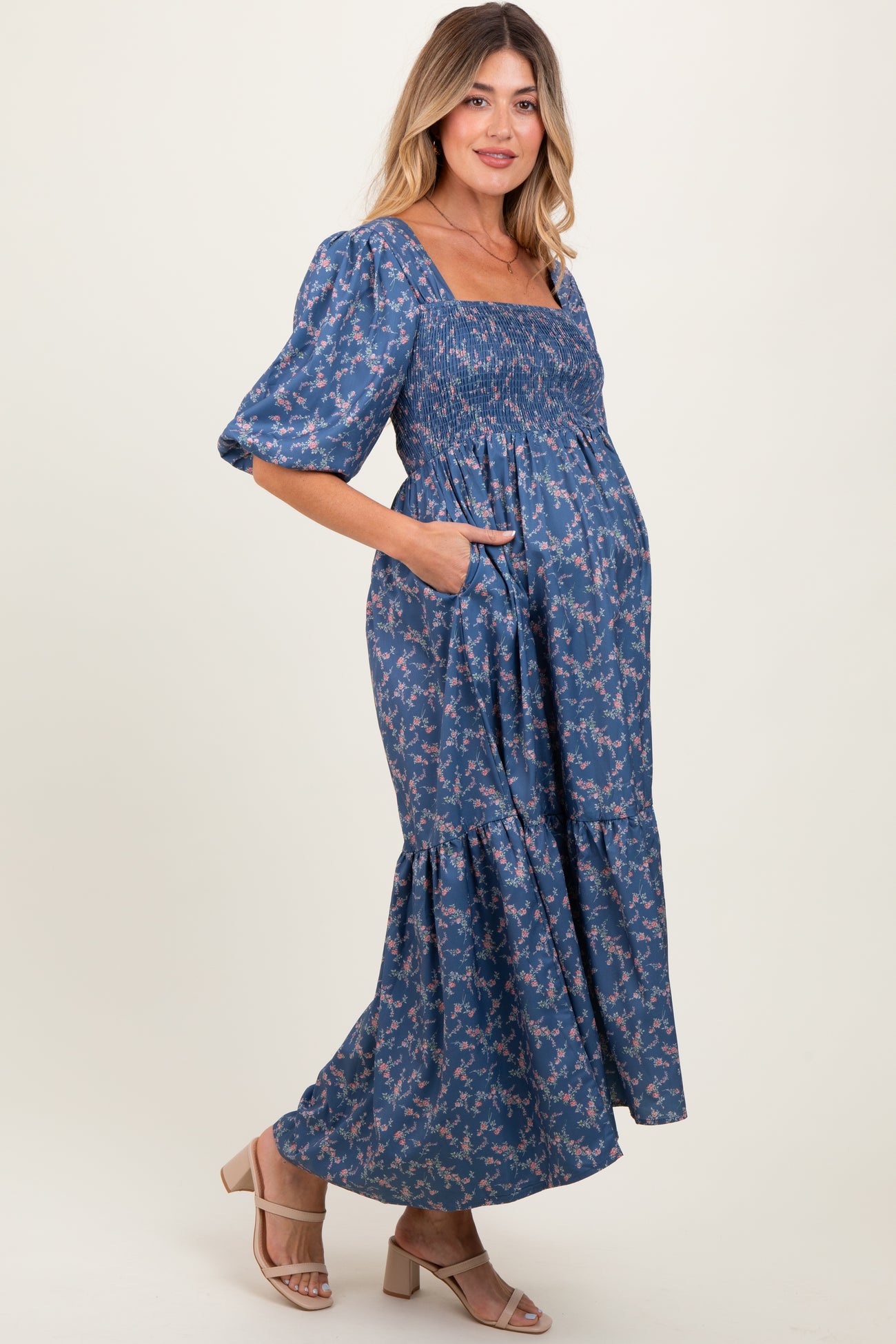 woman wearing a blue floral maternity maxi dress with smocked bodice and puff sleeves, featuring pockets and a baby bump visible