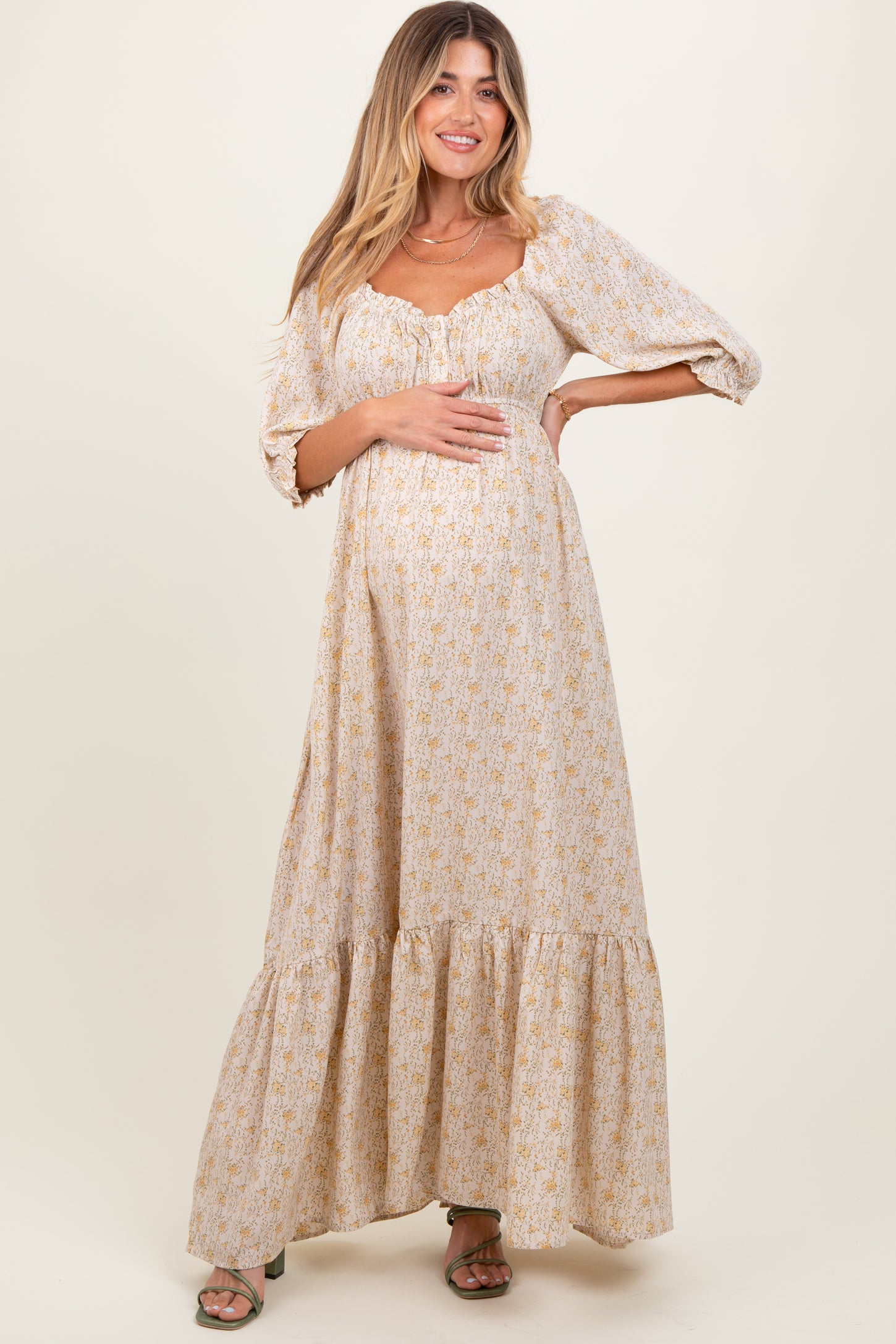 woman wearing a beige floral maternity maxi dress with puff sleeves and a gathered bust, holding her baby bump