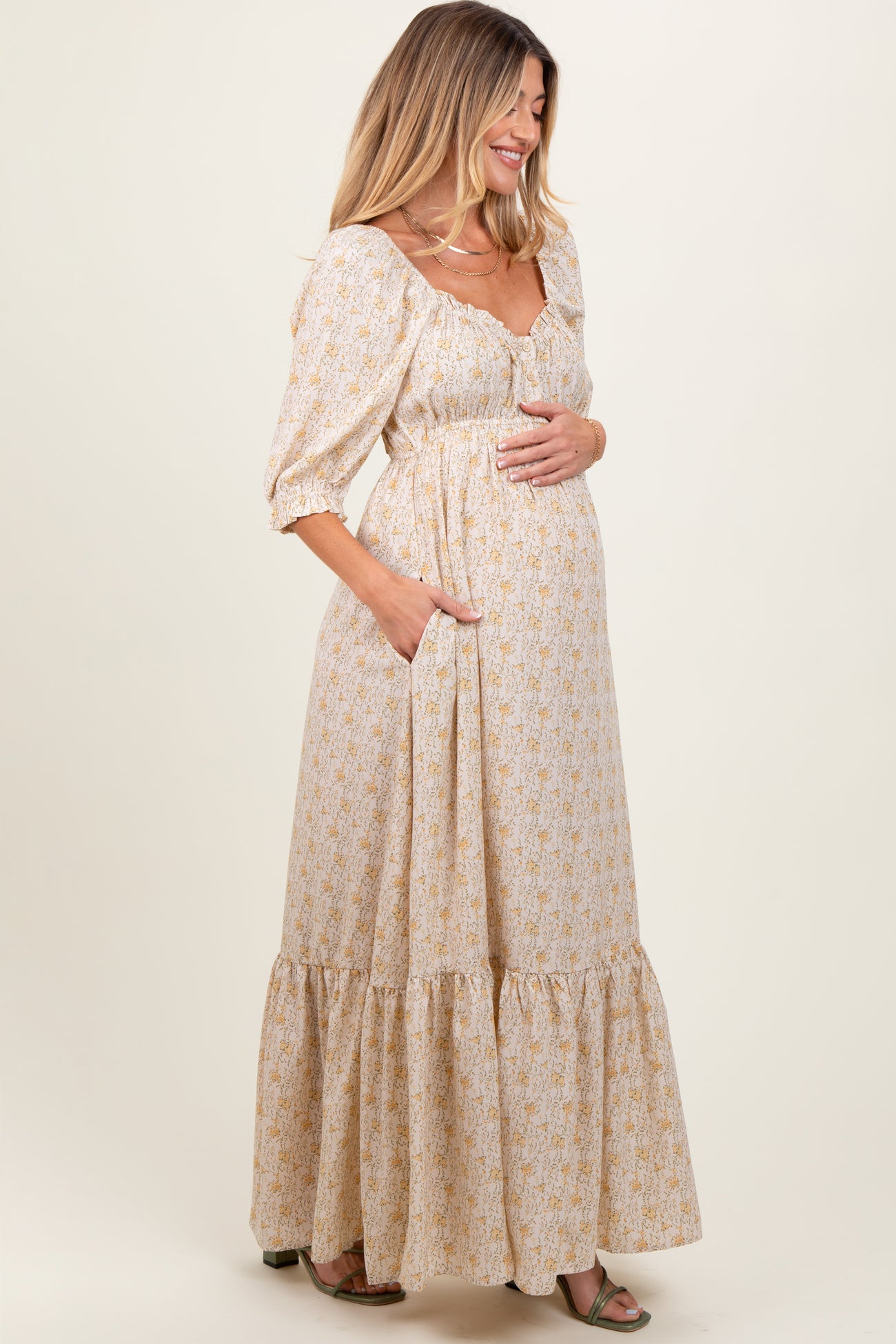 woman wearing a cream floral maternity maxi dress with puff sleeves and pockets, holding her baby bump