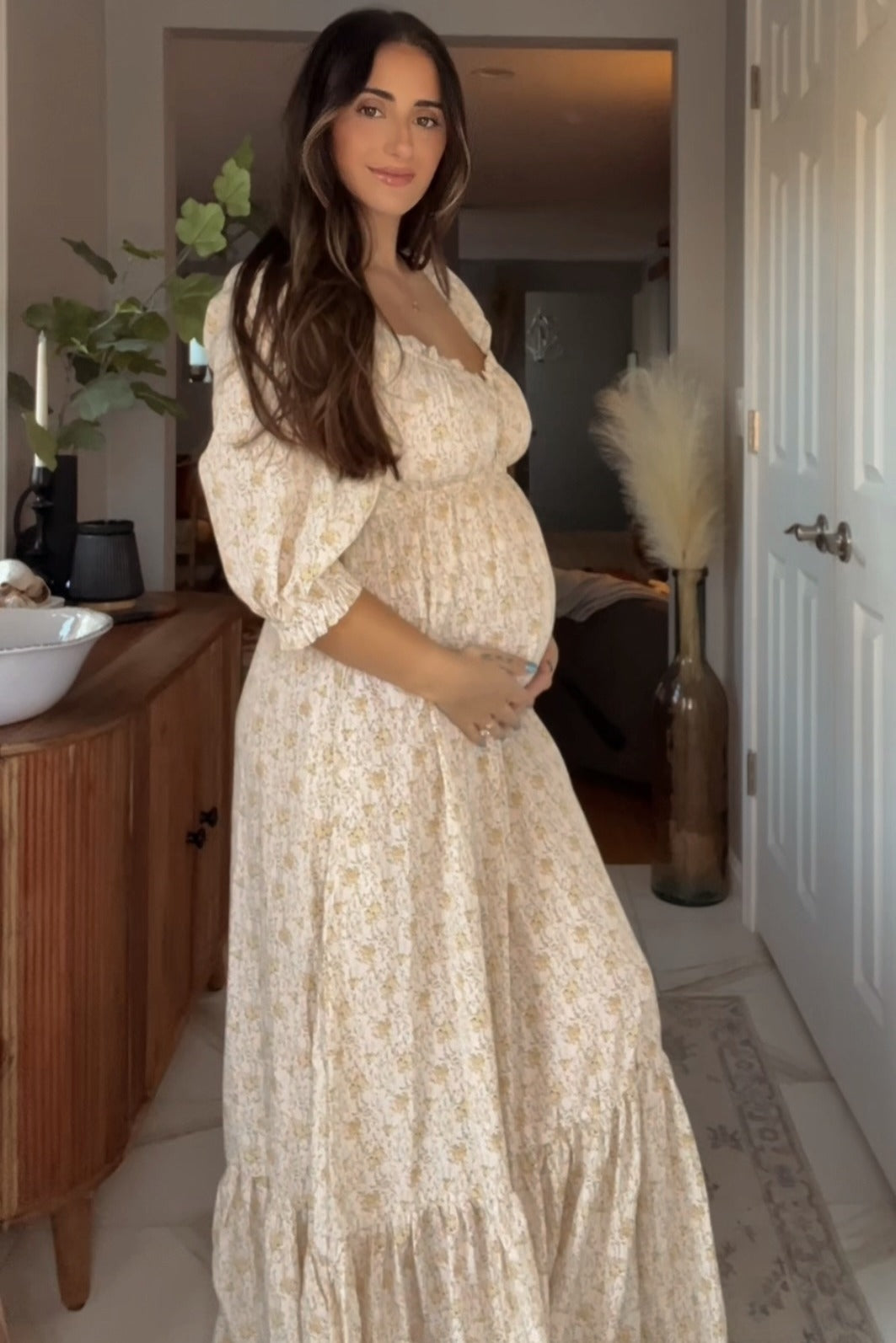 woman wearing a cream floral maternity dress with puff sleeves, holding her baby bump in a sunlit room