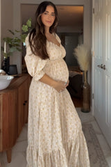 woman wearing a cream floral maternity dress with puff sleeves, holding her baby bump in a sunlit room